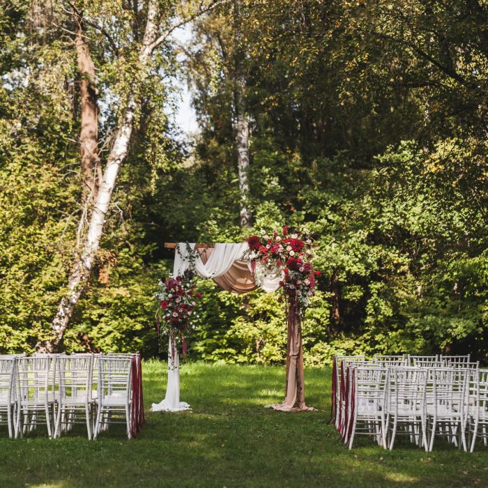 Wedding ceremony in rustic style decorated with different red flowers, white textile and chairs in forest