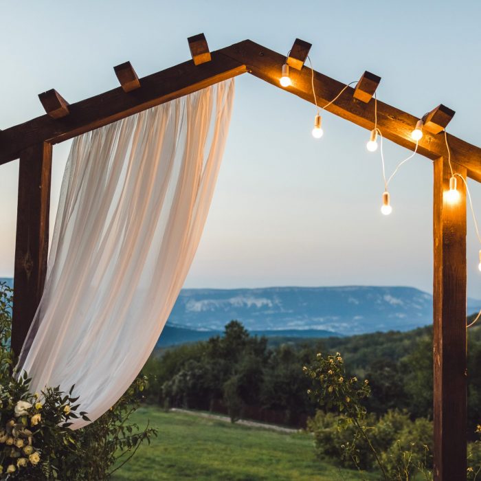 Wooden wedding arch with white cloth and light bulbs outdoors with amazing mountain view on background. Night ceremony in twilight.